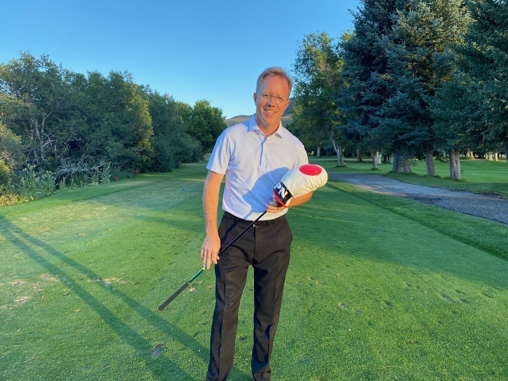 Tom Kotter smiling on a golf course fairway, holding a driver with a head cover
