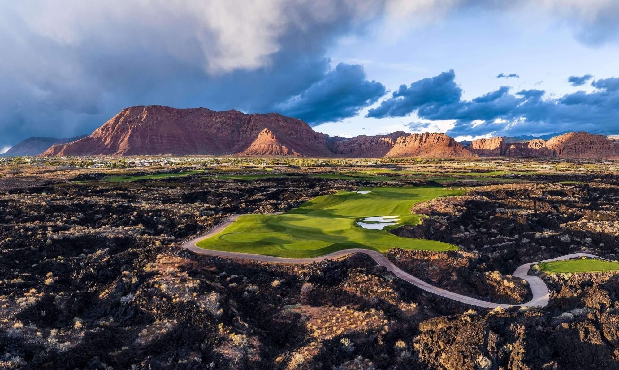 Black Desert Golf Course — vivid green fairway island surrounded by dark lava rock with red sandstone cliffs and stormy sky